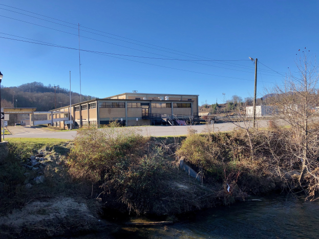 Ein Fluss durchquert eine grüne Wiese neben einem Gebäude in der Nähe von Bäumen, Pflanzen, Gras, Steinen, Straßenlaternen, Strommasten, Stromkabeln, Hügeln und einem klaren blauen Himmel, wobei das Gebäude als Standort einer geplanten Wasseraufbereitungsanlage vorgesehen ist.