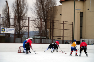 Menschen beim Eishockey auf einer Eisfläche mit Gebäuden, Bäumen, einer Straßenlaterne, einem Namenskennzeichen und Zäunen im Hintergrund unter einem Himmel.