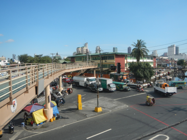 Verkehrsreiche Straße mit Schwerlastverkehr neben einer Brðcke, Fahrzeuge auf der Straße, Fðßler, Zelte, Mäste, Verkehrszeichen, Bäume, Gebäude und Wasser im Hintergrund unter einem bewölktem Himmel.