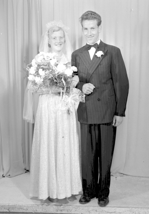 Ein Mann und eine Frau stehen nebeneinander auf einer Bühne, lächeln und halten ein Blumenbouquet in der Hand, mit einem Vorhang im Hintergrund.