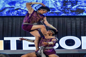 Zwei Performer in purpurfarbenen Kätzen führen Akrobatik auf der Bühne aus, wobei einer den anderen hält, während das Publikum unten zusieht.