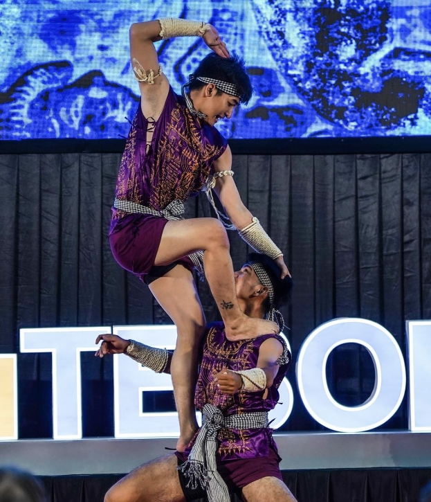 Zwei Performer in purpurfarbenen Kätzen führen Akrobatik auf der Bühne aus, wobei einer den anderen hält, während das Publikum unten zusieht.