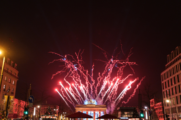 Eine belebte Stadtstraße in Berlin an Silvester, voller Menschen, Fahrzeuge und festlicher Dekorationen, mit Feuerwerk, das den Himmel über Gebäuden und Bäumen erhellt.