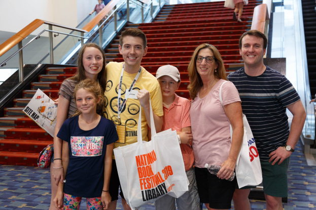Gruppe von Menschen, die vor einer Rolltreppe stehen und Taschen halten, mit Treppen und Geländern im Hintergrund.