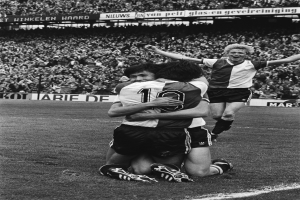 Drei Fußballspieler feiern ein Tor, indem sie sich auf dem Boden umarmen, vor einem jubelnden Publikum und schwenkenden Fahnen im Hintergrund, dargestellt in Schwarz-Weiß.