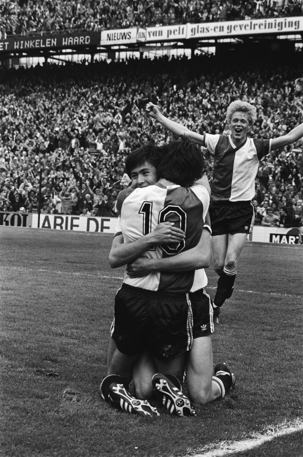 Drei Fußballspieler feiern ein Tor, indem sie sich auf dem Boden umarmen, vor einem jubelnden Publikum und schwenkenden Fahnen im Hintergrund, dargestellt in Schwarz-Weiß.