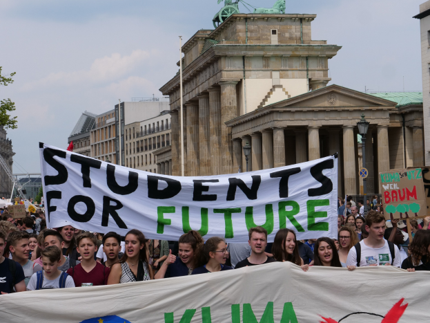 Gruppe von Studenten marschiert in Berlin mit einem bunt bemalten "Students for Future"-Schild vor einer Kulisse aus Gebäuden, Bäumen und Himmel.