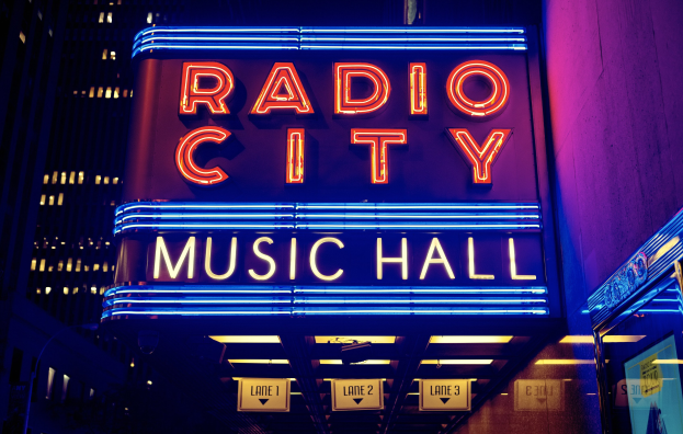 Hell erleuchtetes Radio City Music Hall in New York City mit hohen Gebäuden im Hintergrund, das eine 'Radio City'-Leuchtschrift und mehrere kleinere Schilder an seiner Basis zeigt.