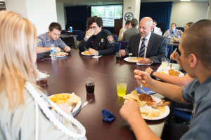 Eine Gruppe von Menschen sitzt an einem Tisch mit Tellern, Gläsern, Besteck und Servietten, mit Säulen, Vorhängen, einem Fenster und Deckenleuchten im Hintergrund.