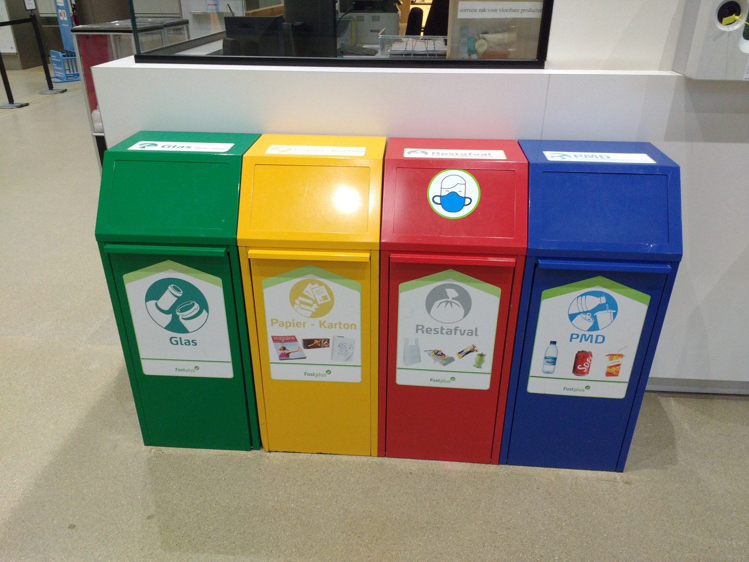 Vier farbenfrohe Mülltonnen in einer Reihe auf dem Boden, mit einer Theke, einem Monitor und anderen Gegenständen im Hintergrund.