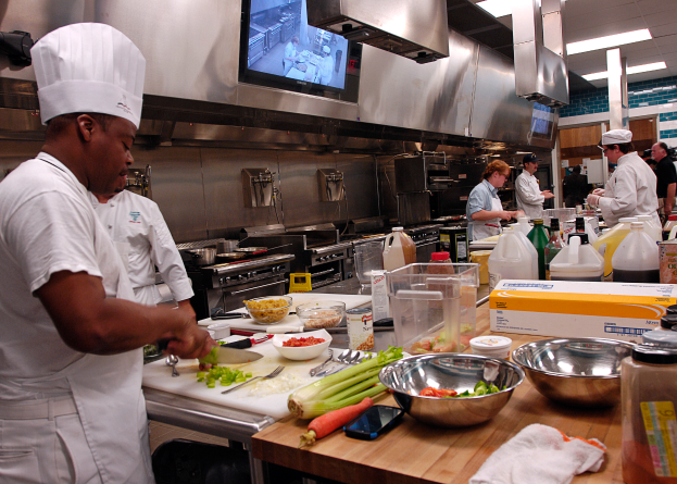 Eine Gruppe von Köchen in einer Küche, die Essen auf einem Schneidbrett zubereiten, mit einem Koch im Vordergrund in einer weißen Schürze und Mütze, der ein Messer hält und von verschiedenen Utensilien, Schüsseln, Gemüse, einem Handy, Flaschen und anderen Gegenständen umgeben ist, mit einem Fernseher und Deckenlampen im Hintergrund.