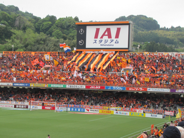 Ein Fußballspiel in einem Stadion mit einer großen Zuschauermenge, saftig grünem Rasen, einem Tor, Bannern, Fahnen, einem großen Bildschirm, Bäumen und einem klaren blauen Himmel.