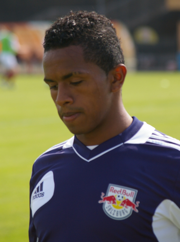 Junger Mann in blauem T-Shirt auf einem Fußballfeld, unscharfer Hintergrund, kurze dunkle Haare, entschlossener Gesichtsausdruck, scheint ein Flügelspieler zu sein.