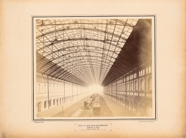 Altes Schwarz-Weiß-Foto eines Bahnhofs mit einer Lokomotive auf den Schienen, flankiert von mehrstöckigen Gebäuden mit Fensterreihen und einem Dach, das von Metallpfosten gestützt wird; Text erscheint unten im Bild.
