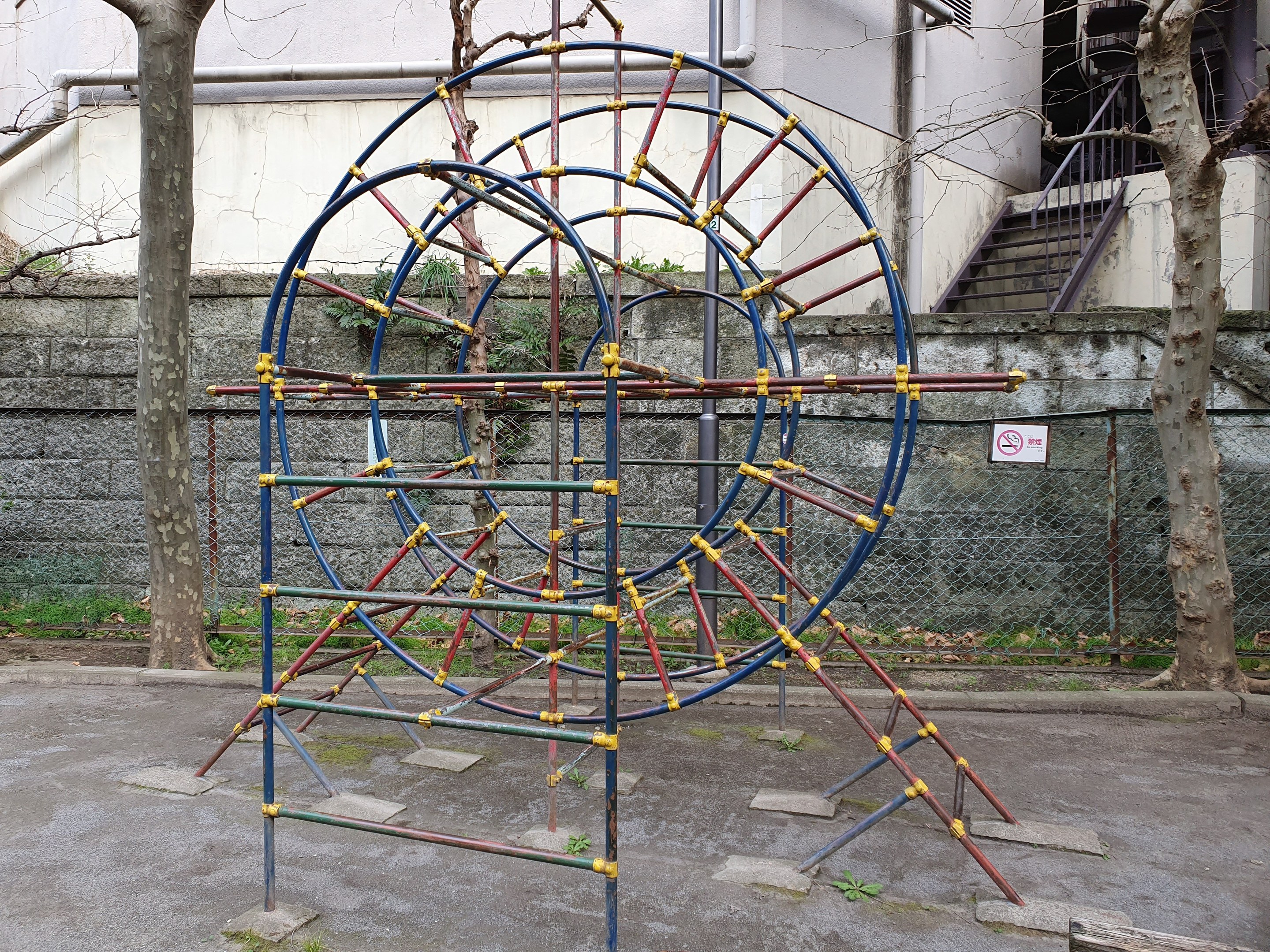 Spielplatz mit großeem Metall-Klettergerüt, umgeben von Bäumen und einer Maschendrahtzaun-Mauer, mit Treppen mit Geländern, die zu einem Gebäude mit Fenstern und Rohren führen, einem Wegweiser und grünem Boden.