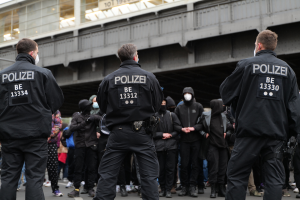 Eine Gruppe von Polizisten in Uniform steht vor einer Menge von Menschen in schwarzen Uniformen und Masken, mit einer Brücke und einem Gebäude im Hintergrund, was eine städtische Umgebung vermuten lässt.
