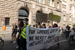 Eine Gruppe von Menschen marschiert auf einer Straße in Berlin, hält Schilder und Banner hoch und fährt mit Fahrrädern, vor einem Gebäude mit Fenstern, Bögen, Säulen, Skulpturen, Bäumen und einem Laternenmast, protestiert gegen die Anti-Kriegsbewegung.