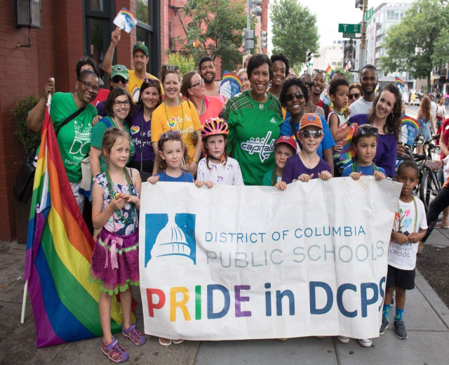 Gruppe von Menschen, die auf dem Boden stehen, einige halten ein Banner mit der Aufschrift "District of Columbia Public Schools Pride in DCPS", mit Kindern, die eine Fahne halten, einem Radfahrer, Bäumen, Gebäuden, Straßenschildern, Ampeln und einem bewölkten Himmel.