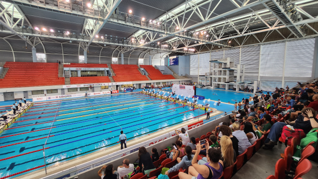 Großer Hallenbad mit vielen Menschen auf Stühlen sitzend und im Wasser stehend, mit Schildern im Hintergrund unter einer Decke aus Lampen und Eisenstangen.
