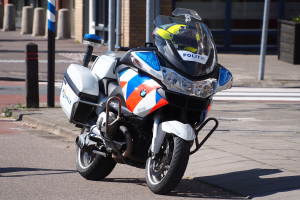 Ein polizeiliches Motorrad am Straßenrand geparkt mit einem Helm darauf, mit Pfählen, Pflanzen und einem Gebäude im Hintergrund.