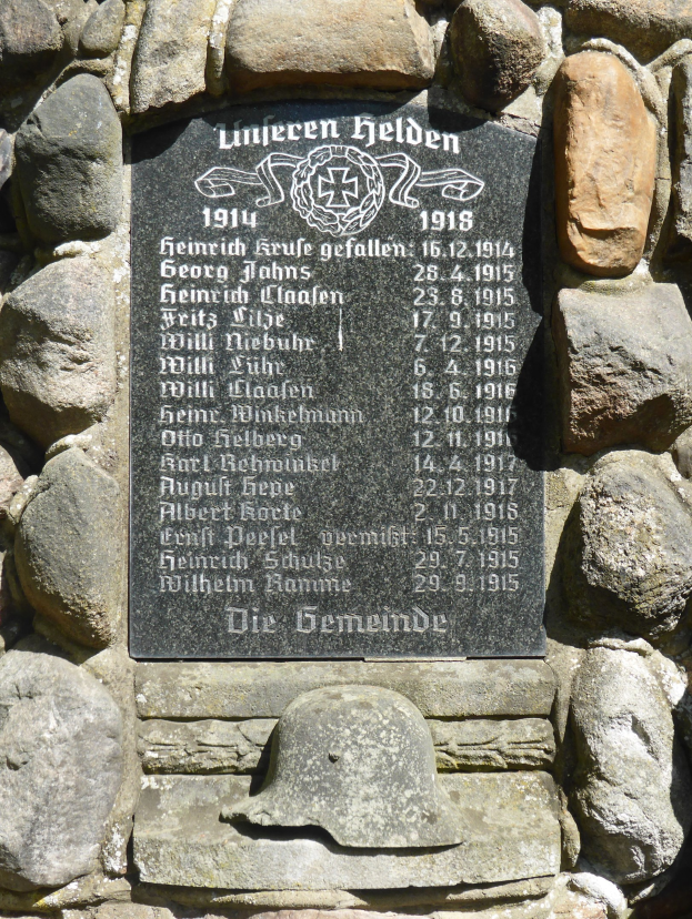 Steinwand mit einer Gedenktafel und einem Helm darauf, beschriftet mit Text.