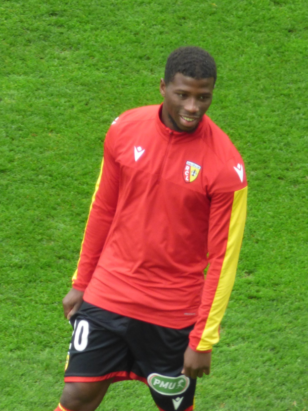 Ein Mann in rotem T-Shirt und schwarzen Shorts auf einem Fußballfeld, der strahlt und eine Flügelposition einnimmt.