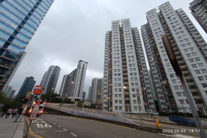 Eine Stadtstraße mit hohen Gebäuden, Fahrzeugen, Fußgängern, Schildern, Laternen, Bäumen und einem bewölkten Himmel, mit einem Wohnhaus im Vordergrund.