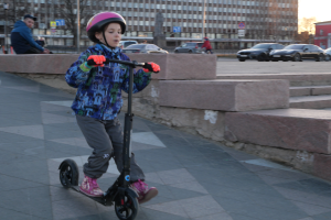 Ein junger Junge fährt mit einem Helm und Handschuhen auf einem Gehweg mit verschiedenen urbanen Elementen und einem klaren blauen Himmel im Hintergrund.