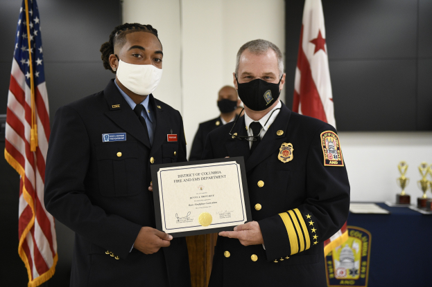 Zwei uniformierte Feuerwehrleute, einer identifiziert als Chef der Feuerwehr des District of Columbia und der andere als stellvertretender Chef, stehen Seite an Seite und tragen Masken und halten ein Zertifikat, mit einem Tisch mit Schilden und Flaggen dahinter.