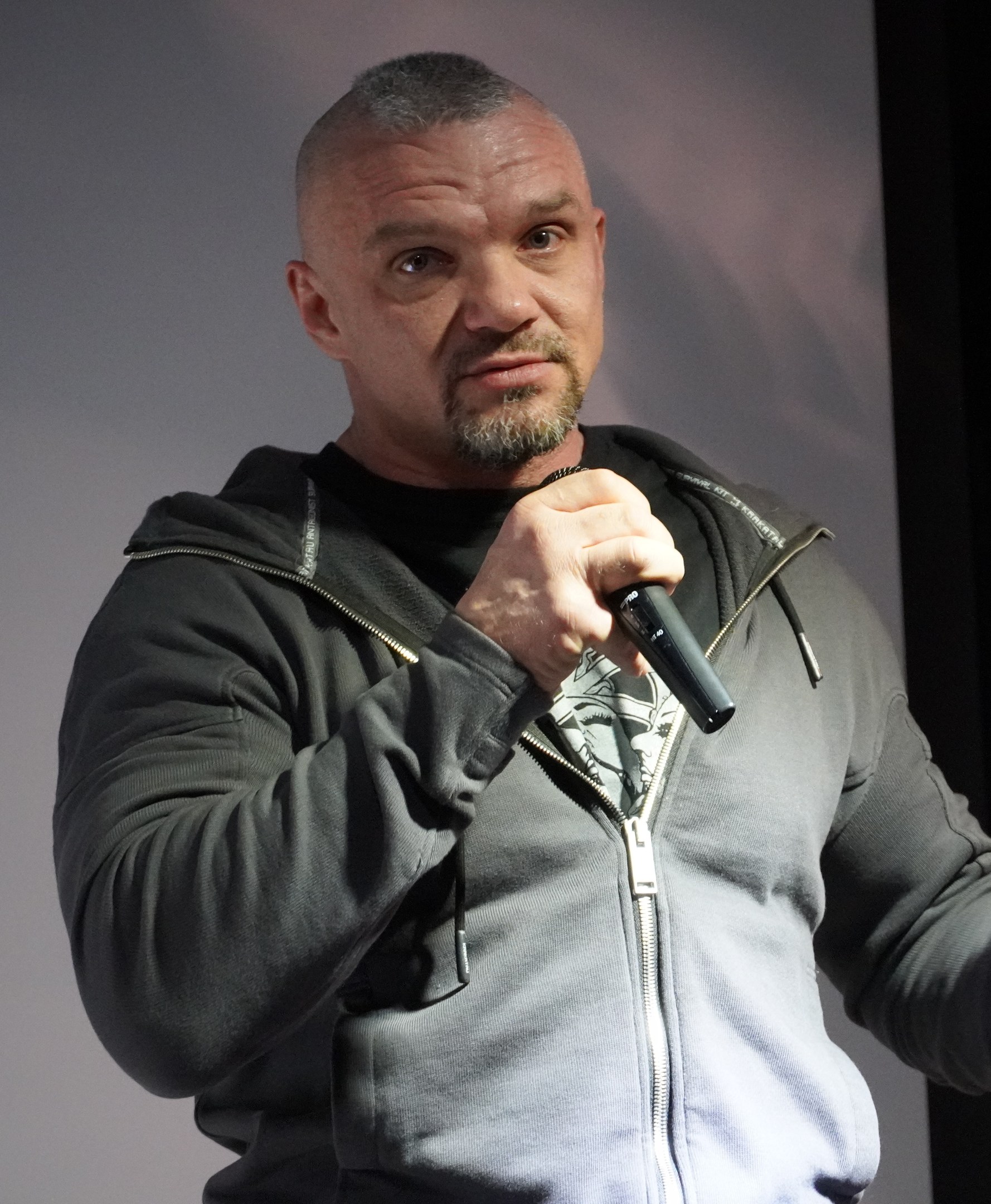 Ein Mann in schwarzer Kapuzenjacke und schwarzem Anzug mit weißem Hemd, der ein Mikrofon hält und selbstbewusst mit zurückgekämmtem Haar vor einer Wand steht.