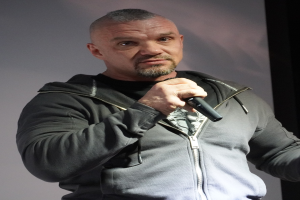 Ein Mann in schwarzer Kapuzenjacke und schwarzem Anzug mit weißem Hemd, der ein Mikrofon hält und selbstbewusst mit zurückgekämmtem Haar vor einer Wand steht.