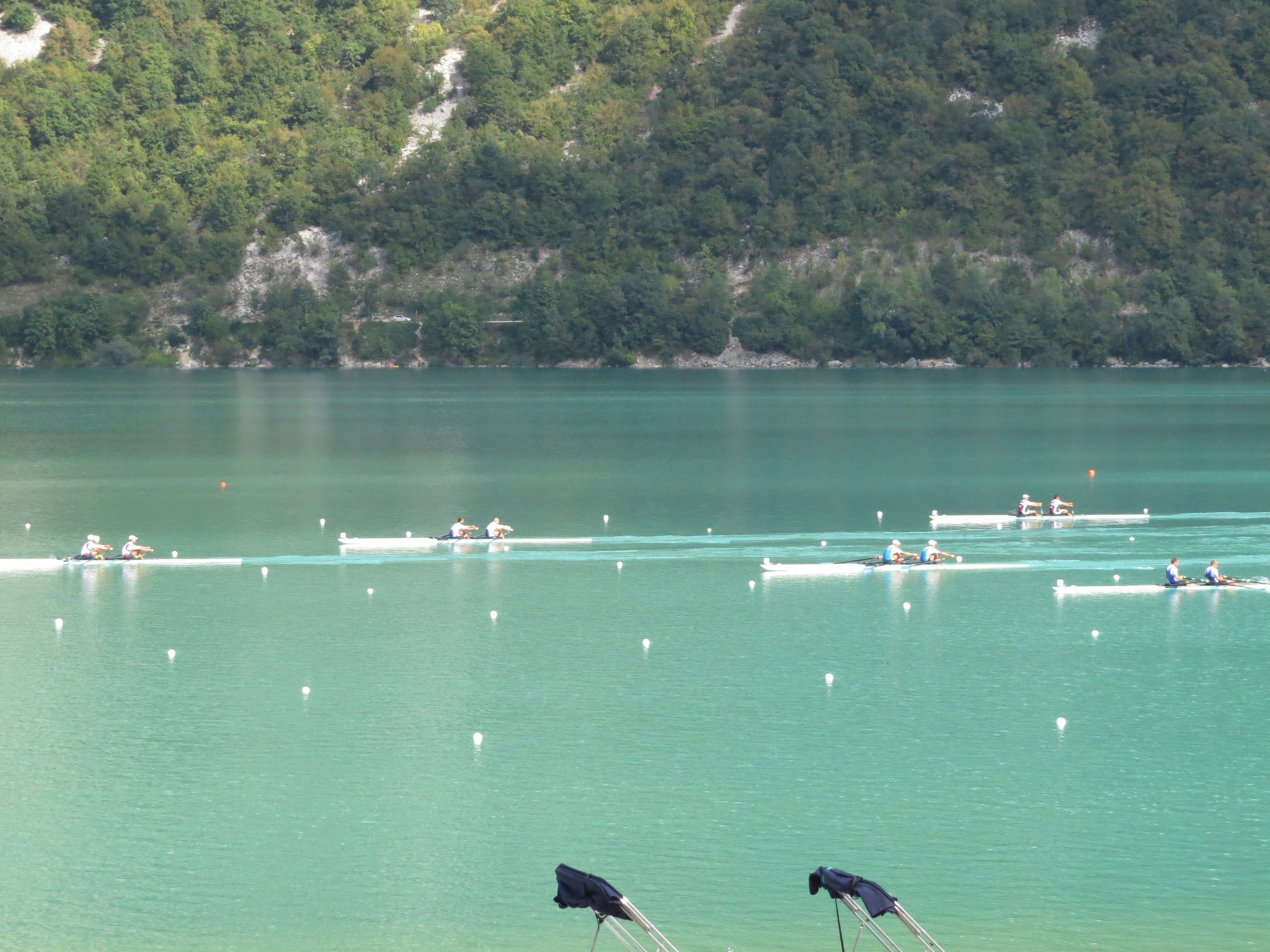 Eine Gruppe von Menschen bei einem Ruderwettbewerb auf einem Gewässer mit Bäumen und einem Hügel im Hintergrund, mit Kleidung über den Tribünen am Boden.