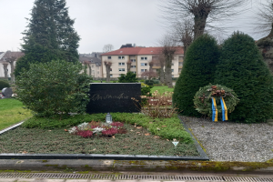 Ein Mahnmal für die Opfer des Zweiten Weltkriegs steht in einem Park, geschmückt mit einem Kranz und umgeben von Grün, mit Gebäuden und Himmel im Hintergrund.
