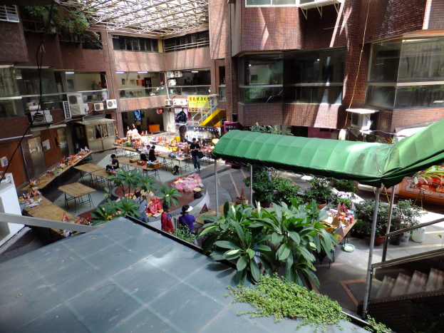 Großer Atrium mit üppiger Vegetation, Menschen an Tischen mit Gegenständen, Treppen mit Geländern und Gebäude mit Fenstern und Klimaanlagen im Hintergrund.