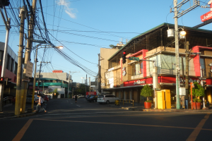 Eine Stadtstraße mit Autos, Fußgängern, Strommasten, Ampeln, Gebäuden mit Schildern, Topfpflanzen und einer bewölkten Himmel.