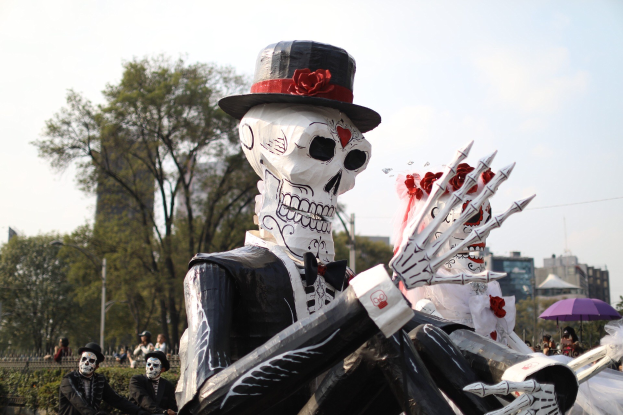 Ein Day of the Dead-Umzug in Mexiko-Stadt mit Menschen in Kostûmen mit Schirmen, skelettartigen Figuren, Bâumen, Gebâuden, Laternenmâsten und einem bewölkten Himmel, mit Text unten.