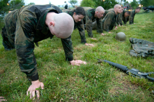 Eine Gruppe von Männern in Militäruniformen, die Liegestütze auf dem Gras machen, mit einer Waffe, einem Helm und anderen Gegenständen, die herumliegen, unter einem klaren blauen Himmel mit Bäumen, Lichtern und Fahrzeugen im Hintergrund.