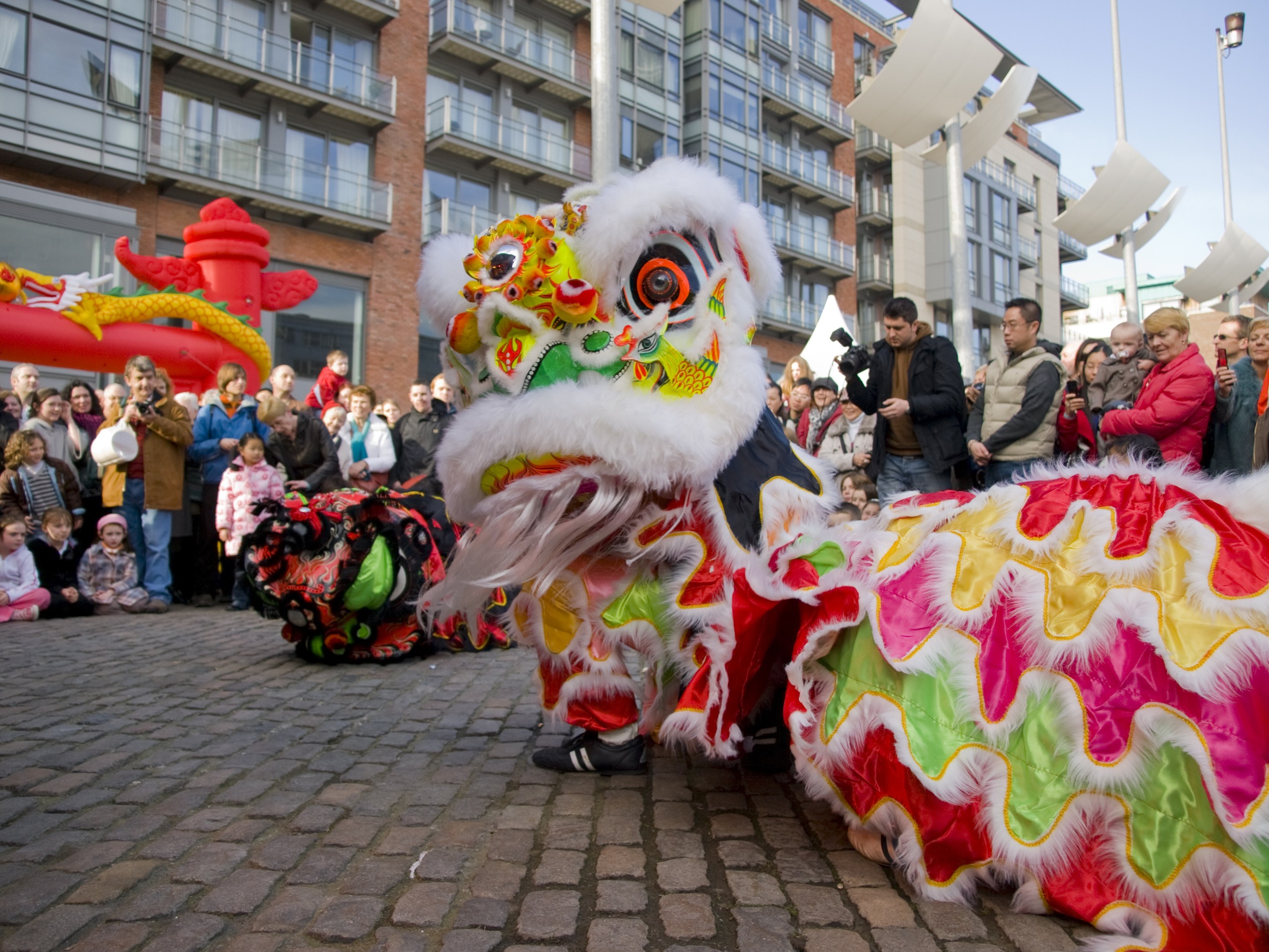 Ein lebendiges chinesisches Neujahrsfest in Amsterdam mit einem Löwen tanzen im Vordergrund und einer Menschenmenge, die Kameras hält, die sich darum versammelt hat, vor einem Hintergrund aus Gebäuden, Laternenmasten und einem klaren blauen Himmel.