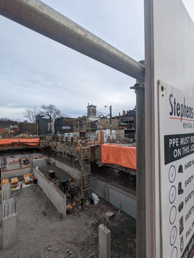 Baustelle mit einem Gebäude-Schild, auf dem "Stephen's Rental Services" steht, und einer Tafel auf der rechten Seite, die Gebäude, Bäume, Versorgungsmasten und einen klaren blauen Himmel im Hintergrund zeigt.