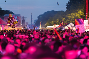 Eine große Menschenmenge auf einem nächtlichen Musikfestival, beleuchtet von bunten Lichtern, mit Zelten, Fahnen, Masten und anderen Gegenständen drumherum und einem Turm, Bäumen und einem klaren Nachthimmel im Hintergrund.