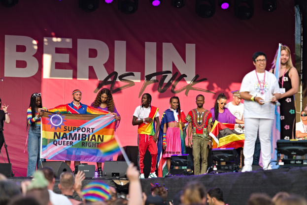 Gruppe von Menschen auf einer Bühne mit einem Banner, auf dem 'Berlin as Fuck' steht, mit Lautsprechern, Mikrofonstativen und einer feiernden Menge unten, die Hände hebt.