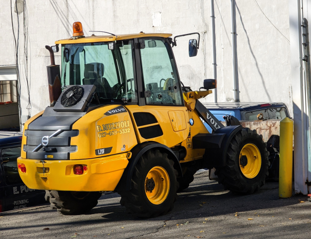 Ein gelber Radlader auf einer Straße vor einem Gebäude mit einer Wand im Hintergrund, mit anderen Fahrzeugen in der Nähe.
