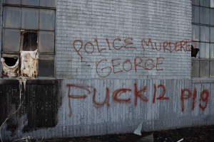 Graffiti an einer Gebäudewand mit der Aufschrift "Polizei Mord George Fuck 12 P19" mit Fenstern und verstreuten Gegenständen davor.