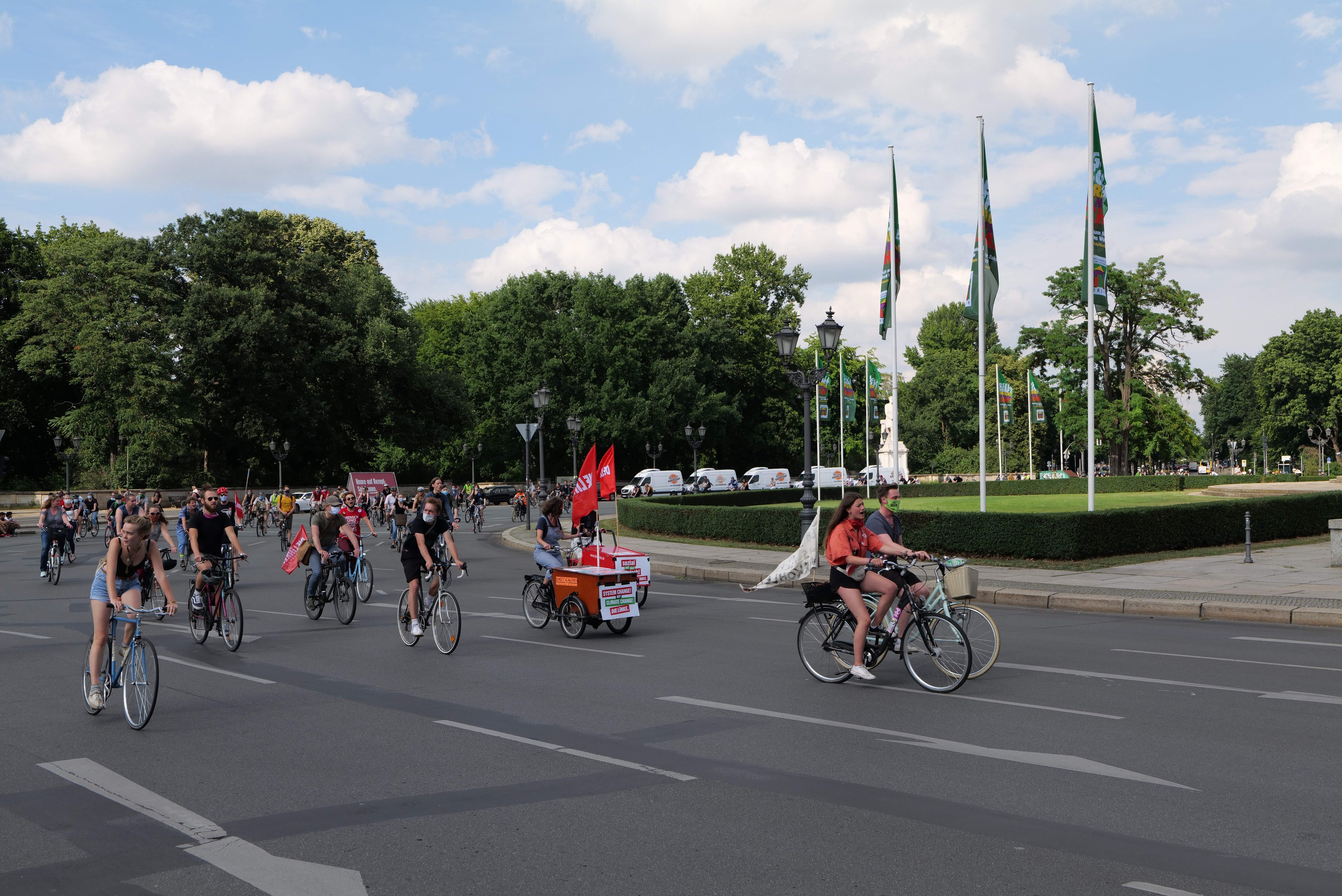 Eine Gruppe von Radfahrern fährt eine von Bäumen gesäumte Straße mit Laternenmasten und Fahnenmasten während der Pariser Radfahrt 2016, mit bewölktem Himmel und Fahrzeugen im Hintergrund.