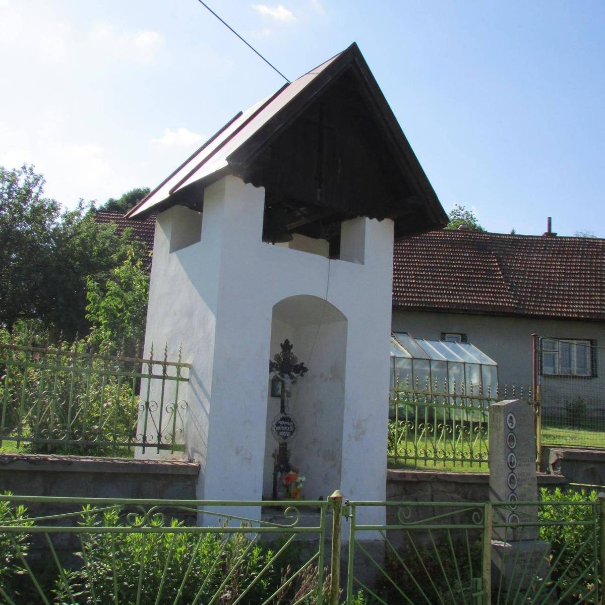 Kleine weiße Kirche mit einem Kreuz darauf, umgeben von einem Metallzaun, Pflanzen und einem Pfad, vor Häusern und einem bewölkten Himmel.