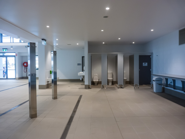Große öffentliche Toilette mit mehreren Toiletten, tragenden Säulen, einem Müllbehälter, einer Ausgangstür, einer Hinweistafel, einem Feuerlöscher und Deckenleuchten.