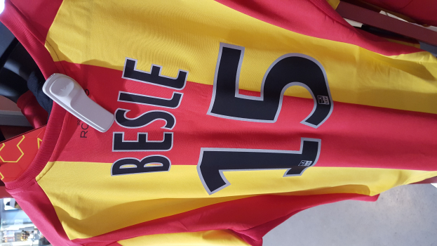 Rotes und gelbes Fußballtrikot mit aufgedrucktem Text und Nummern an einem Ladengestell hängend, mit anderen Gegenständen und dem Boden im Hintergrund.