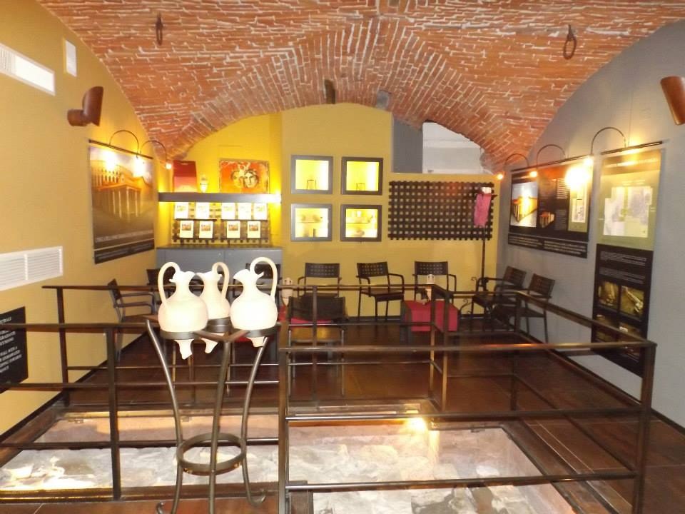 Ein geräumiger Museumsraum mit mehreren Tischen, Stühlen, Displaytafeln, gerahmten Kunstwerken an den Wänden, Vasen, Leuchten und anderen Ausstellungsstücken.