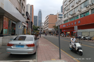 Ein Mann fährt mit einem Roller die Straße zwischen hohen Gebäuden entlang, mit Menschen auf den Bürgersteigen und Fahrzeugen auf der Straße, unter einem klaren blauen Himmel mit Bäumen im Hintergrund.