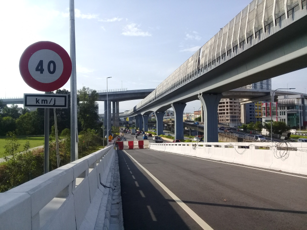 Autobahn mit Tempolimit-Schild, mehreren Fahrzeugen, Brücke mit Pfeilern, Laternen, Bäumen, Gebäuden und bewölktem Himmel im Hintergrund.